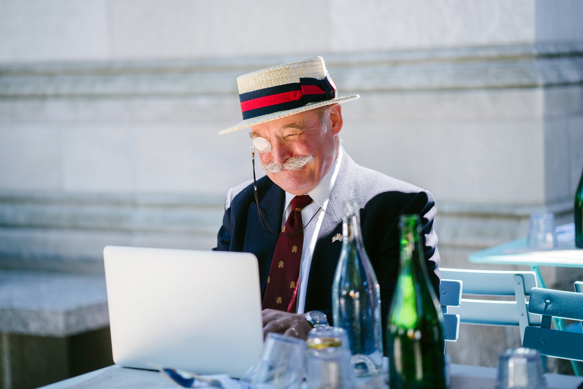 man using laptop at outdoor cafe wearing a hat and a monocle